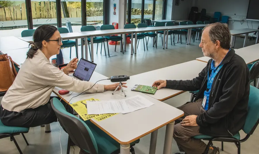 Two people sit at a desk with a laptop and microphone to record the interview