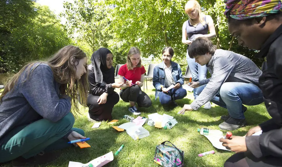 a group of young poeple in a circle on the grass, looking over papers and post-it notes