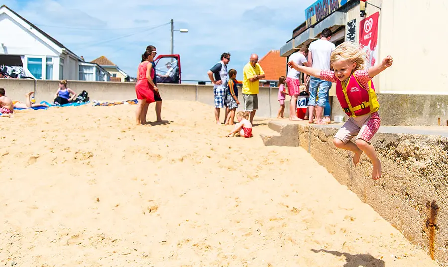 A young girl jumping from a wall onto the sand at Jaywick in Essex. Families are in the background at a cafe.