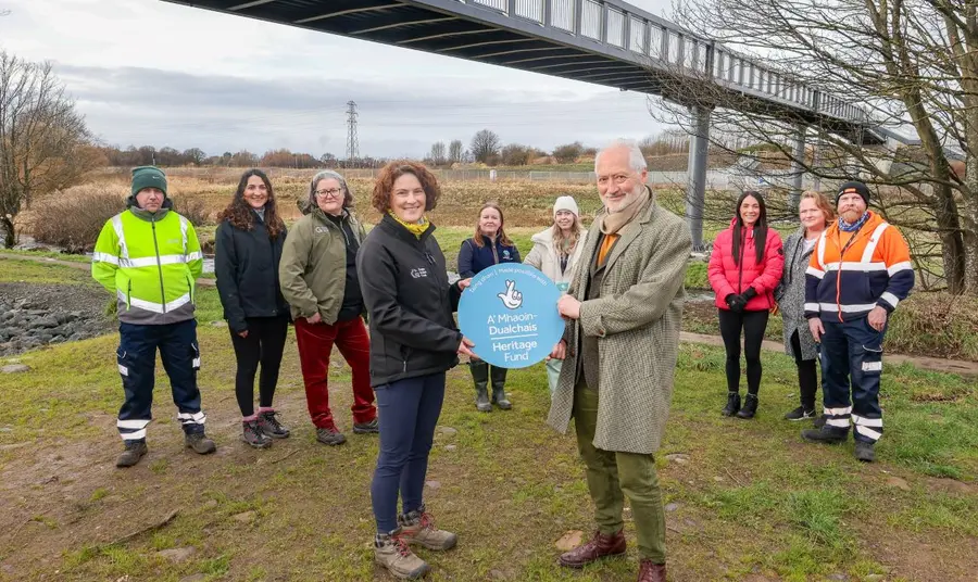 A group of representatives from different organisations involved in the River Park project gather together on the banks of the river, with a pedestrian bridge crossing behind them. To the front, two hold a Heritage Fund roundel.