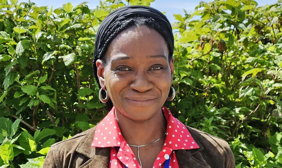 A head and shoulders portrait of a black woman standing in front of garden bushes