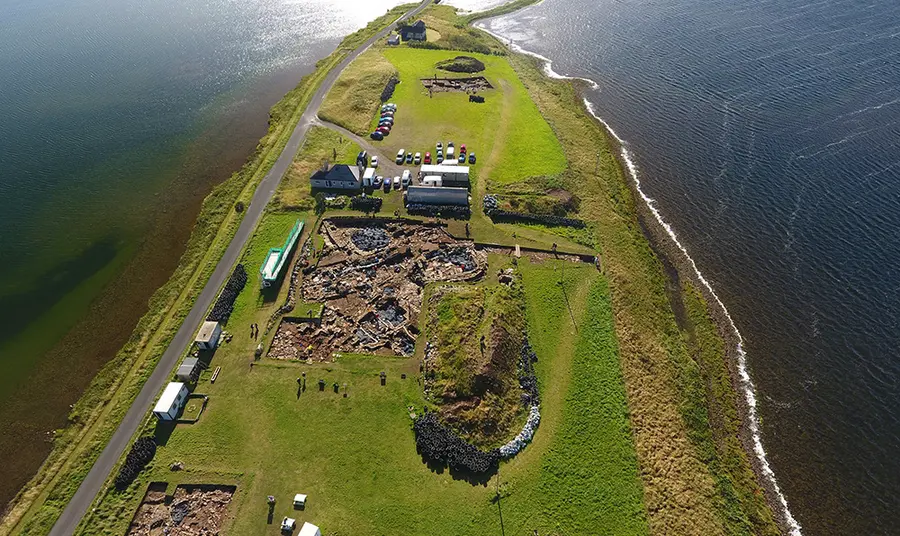 Looking South East over the site, a finger of grass covered land with water either side and a thin causeway visible at the top of the image. Trench J in bottom left; Trench P in centre, and Trench T in distance
