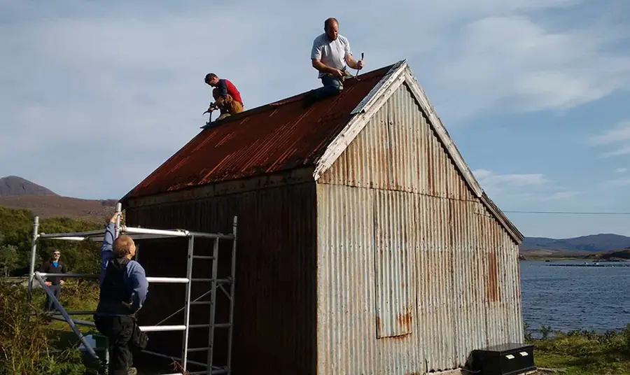 Men working on a small corrugated building with water and mountains in the distance.