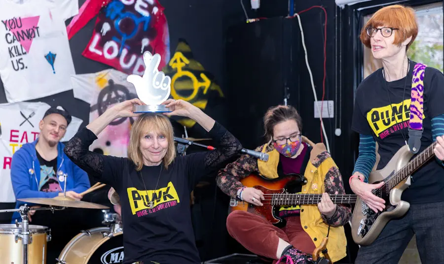 Four people play musical instruments including the drums and guitar in front of memorabilia from the Punk era