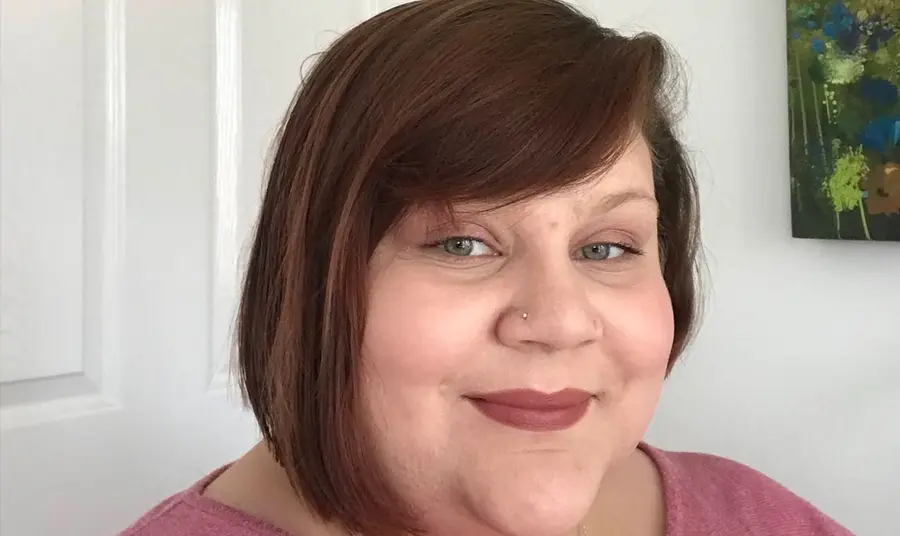 A selfie of a women with shoulder-length brown hair, standing against a white wall