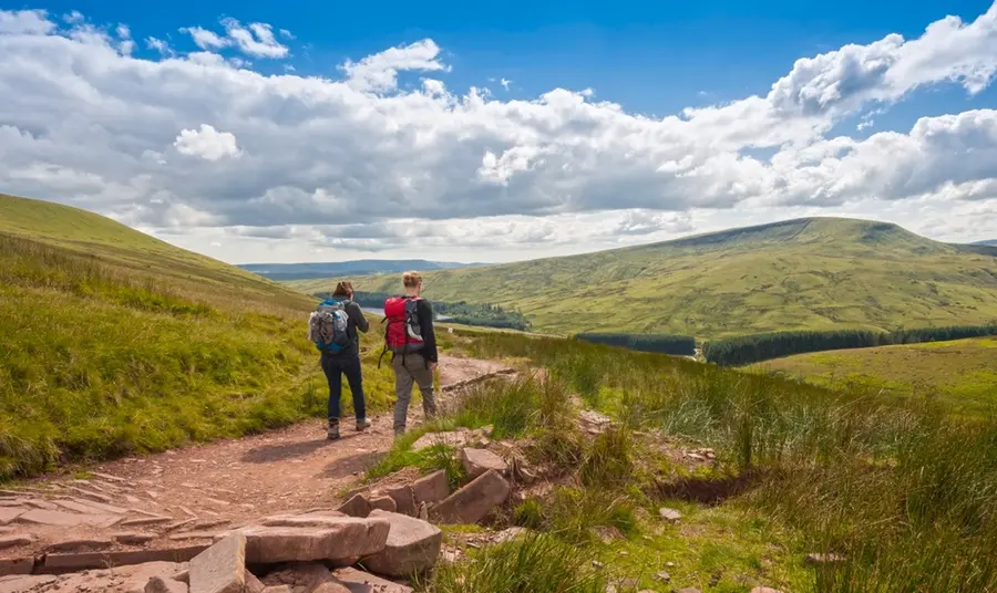 Two hikers walk away down a dirt track into an open, green expanse of the Brecon Beacons, on a sunny day.