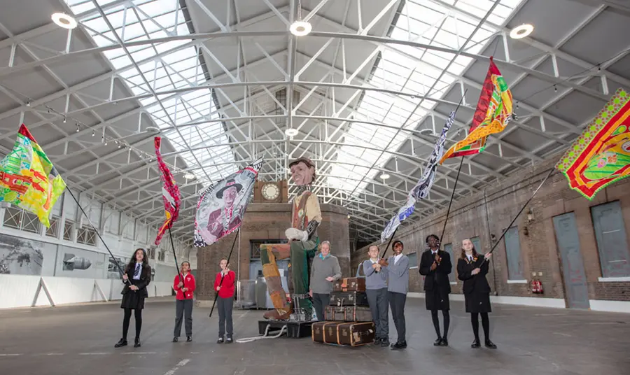 A couple of school children holding large flags in an empty building, accompanied by a giant puppet