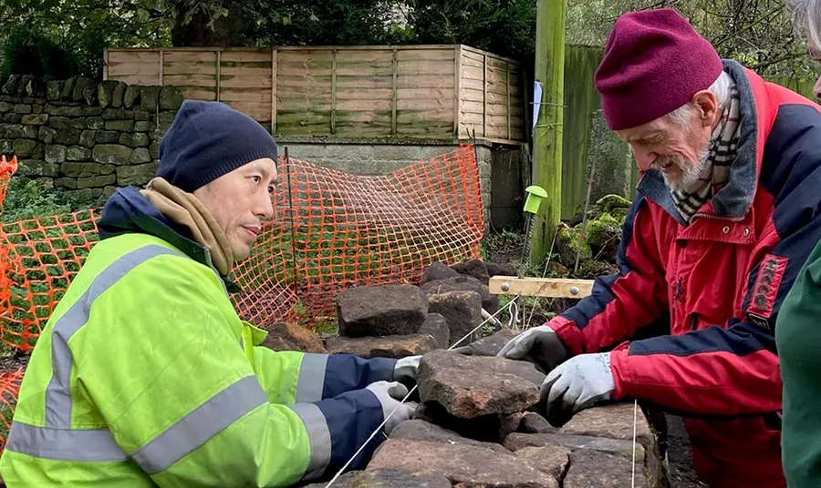 A person demonstrates dry stone wall construction techniques to two people in an outdoor setting