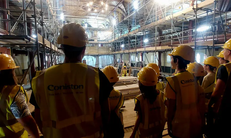 a group of people wearing hard hats and high-vis jackets stand inside the building looking at conservation works