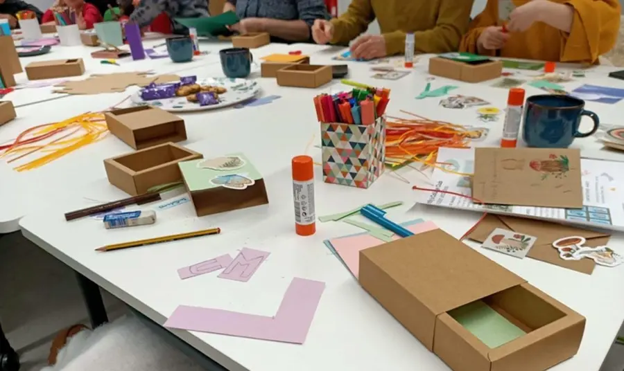 people sit around a large table filled with craft materials, tea and biscuits for a creative workshop