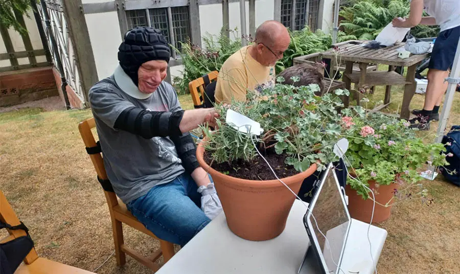 A smiling person sits on a chair and another person crouches down and holds their hand. Together they touch the leaves of a potted plant on a table in front of them. Wires run from the plant to a tablet.