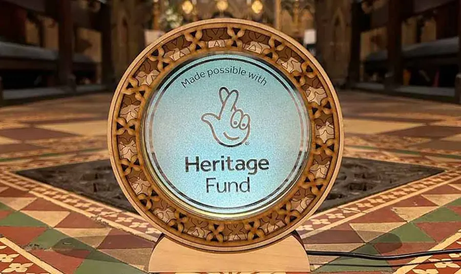 The National Lottery Heritage Fund acknowledgement stamp etched onto a circular glass plaque with a fretwork boarder. The plaque sits in front of a colourful tiled floor and an ironwork grate.