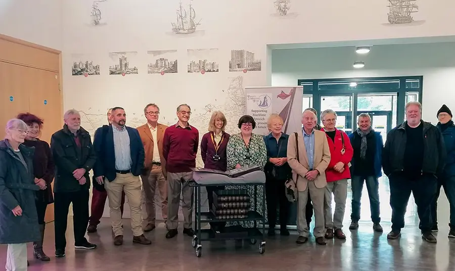 a group of 15 people pose for a photograph, standing behind a historic postcard album on a trolley