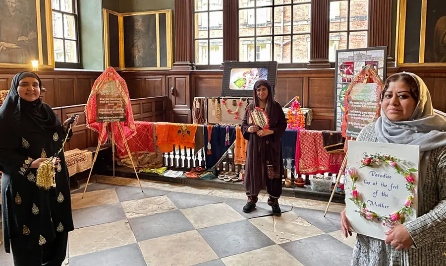 Three Pakistani women holding personal items, standing in front of an exhibition in a small chapel