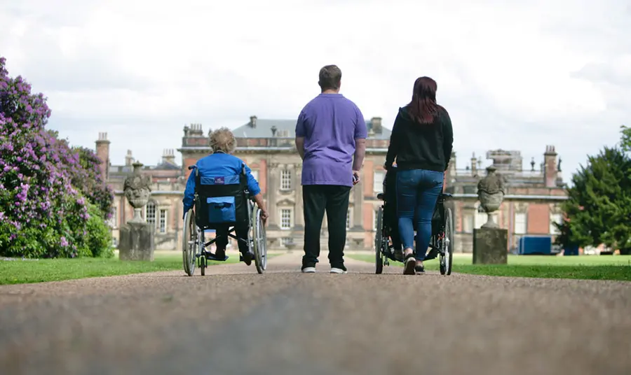 Two people in wheelchairs, and two others walking beside them, travel down a long path towards a stately home.