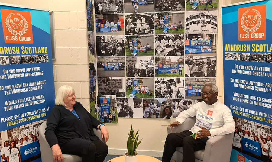 Two people talking. Behind them are banners promoting the Windrush Scotland project.