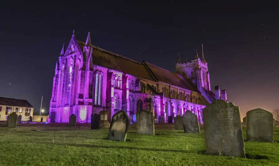 at night a large church is lit up using coloured spotlights. in the foreground are gravestones