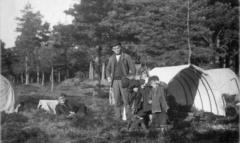 A historic black and white photograph of traveller children and an adult standing in front of tents