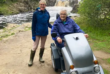 A woman on a mobility scooter, with another woman standing beside her, in an outdoor landscape. A waterfall can be seen in the background.