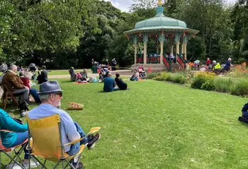 People sitting on camping chairs and on the grass in Pearson park in front of a historic bandstand
