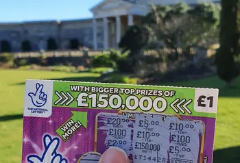 A National Lottery scratchcard held in front of a large building with classical architecture and lawn 