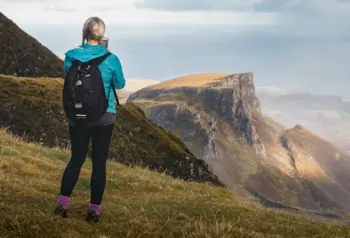 A person standing on a grassy mountainside, looking out to mountains in the distance