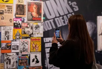 Image of person with long dark hair and dark clothing taking a picture on a mobile of a display of music gig posters at The Museum of Youth Culture