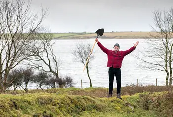Person standing in nature looking happy whilst holding a spade in the air