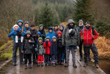 A large group of people of all ages in walking gear on a rainy day. They are standing on a path through a wooded glen.