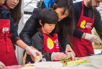 A child in an apron making kimchi with the help of an adult