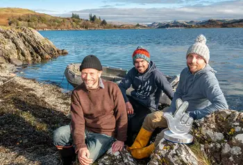 Three people sit on the edge of a loch in Scotland, with a boat in the background and the award in front of them