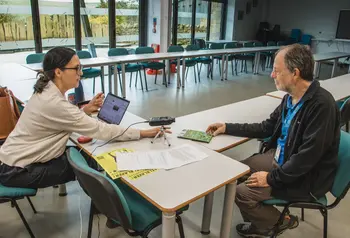 Two people sit at a desk with a laptop and microphone to record the interview