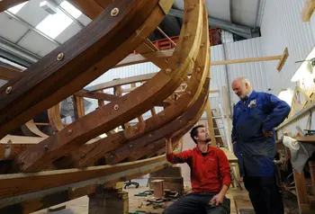 An older man stands alongside the wooden frame of a boat. A younger man is sitting next to the boat with his hand on the frame.