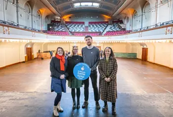 A group of four colleagues, holding up a blue Heritage Fund roundel, stand in the middle of the stage of the Leith Theatre, looking out to the empty stalls and grand circle above.