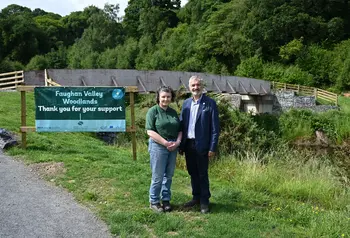 Two people are stood in front of the new bridge in Faughan Valley woodlands. Next to them is a sign to introduce the woodlands and has the Heritage Fund logo on it.