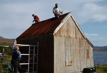 Men working on a small corrugated building with water and mountains in the distance.