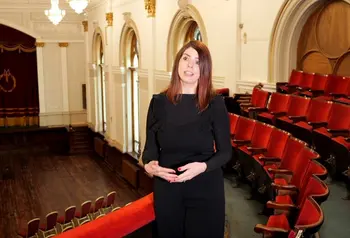 A woman in a black dress standing in an empty theatre speaking to the camera. Red chairs and a curtained stage can be seen in the background.