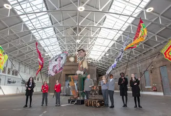 A couple of school children holding large flags in an empty building, accompanied by a giant puppet