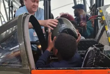 Photograph of a young person sitting in the cockpit of a plane. Someone is passing him a helmet.