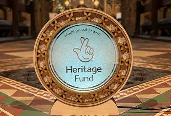 The National Lottery Heritage Fund acknowledgement stamp etched onto a circular glass plaque with a fretwork boarder. The plaque sits in front of a colourful tiled floor and an ironwork grate.