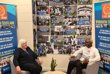 Two people talking. Behind them are banners promoting the Windrush Scotland project.