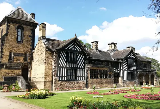 A large brick building with some Tudor-style features. In the foreground are gravel paths and a lawn with flowers planted in it.