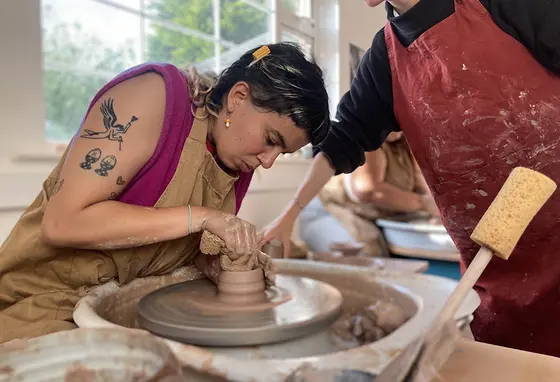 A person in an apron helping another person use a pottery wheel