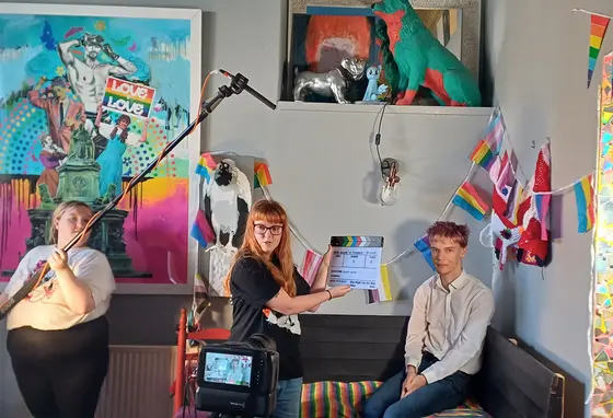 Three young people making a film, one person is holding a microphone, another a clapperboard, and one person is waiting to be interviewed