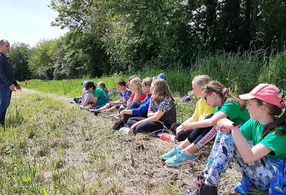 A standing man talking to children sitting on the edge of woodland