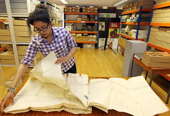 A person handles large parchment documents in an archive room, with shelves of archive boxes in the background