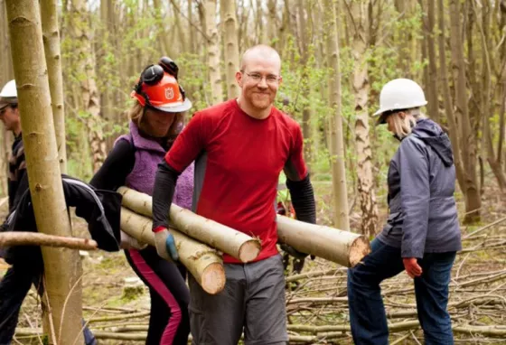 Two people carrying three cut tree trunks through a woodland.