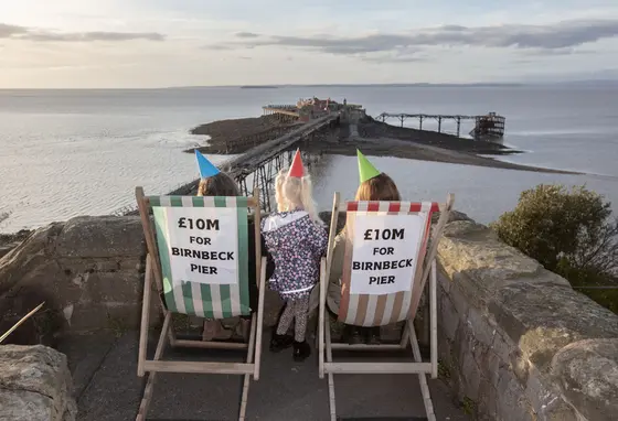 People sitting on deck chairs wearing party hats in front of Birnbeck Pier