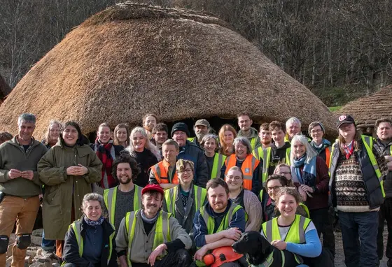 A group of people some wearing high-vis jackets stand together in front of a round house made from wood with a thatch roof