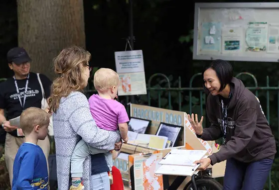 A woman stands outside in front of a bike-mounted display of pictures and other exhibits. She holds a clipboard and talks to a woman and two young children.